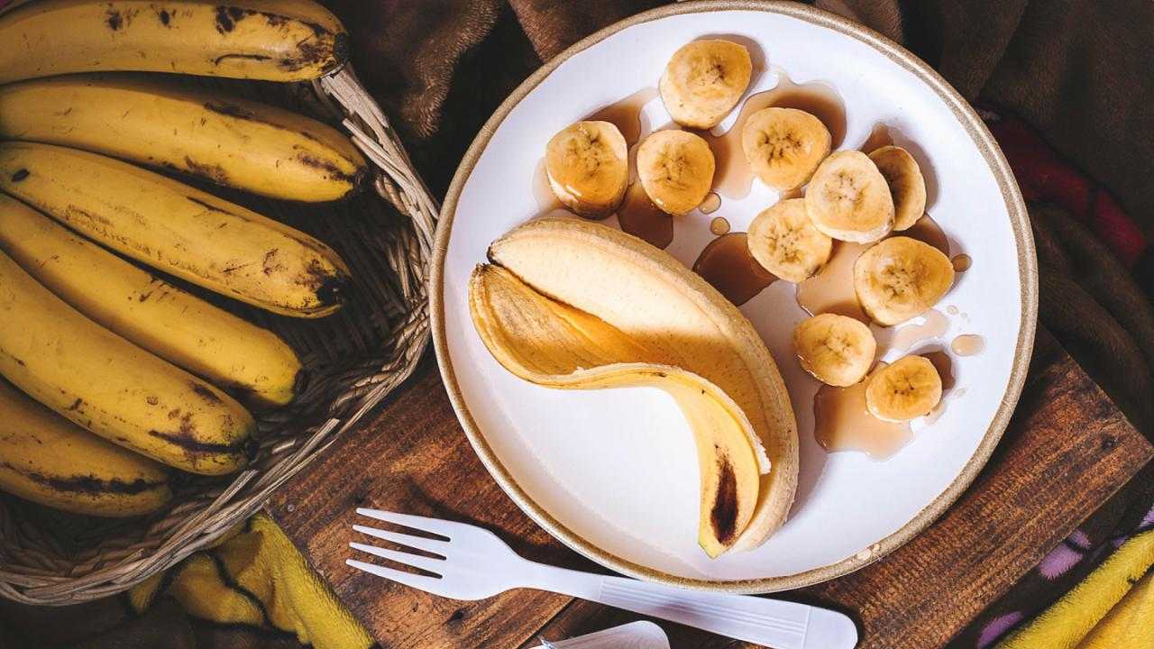 Bananen enthalten viel Kalium und sollten bei Dialyse lieber gemieden werden. Banane und Bananenstücke liegen auf einem weißen Teller neben einem Bananenkorb